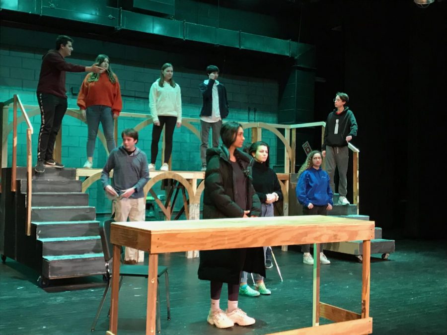 Theater Company members stand solemnly on stage at the beginning of a scene in an after school rehearsal.